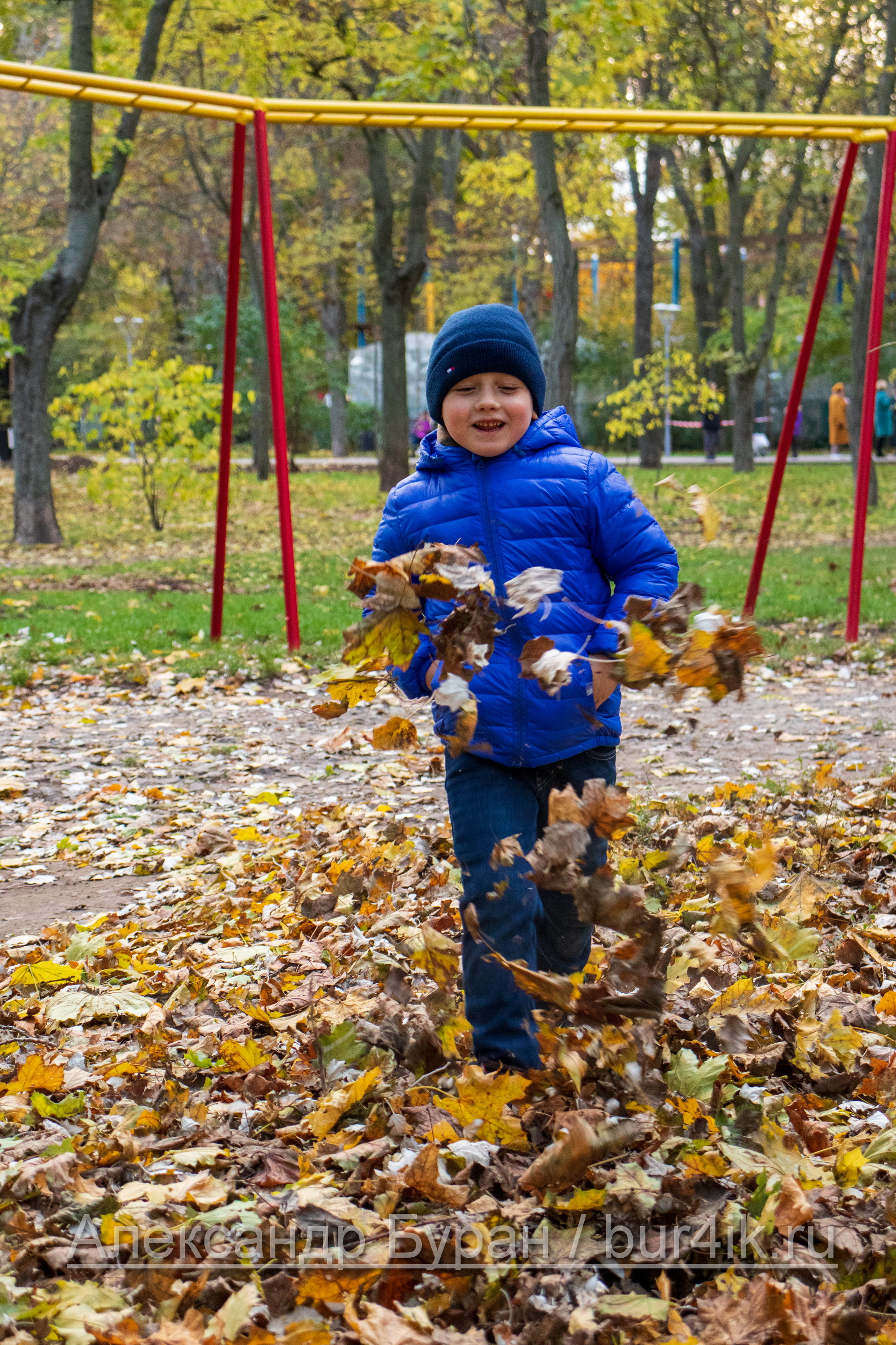 Мальчик в синей куртке подбрасывает желтые листья ногами в парке осенью