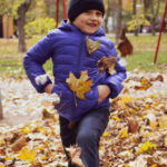 Мальчик играет в синей куртке с листьями в осеннем парке