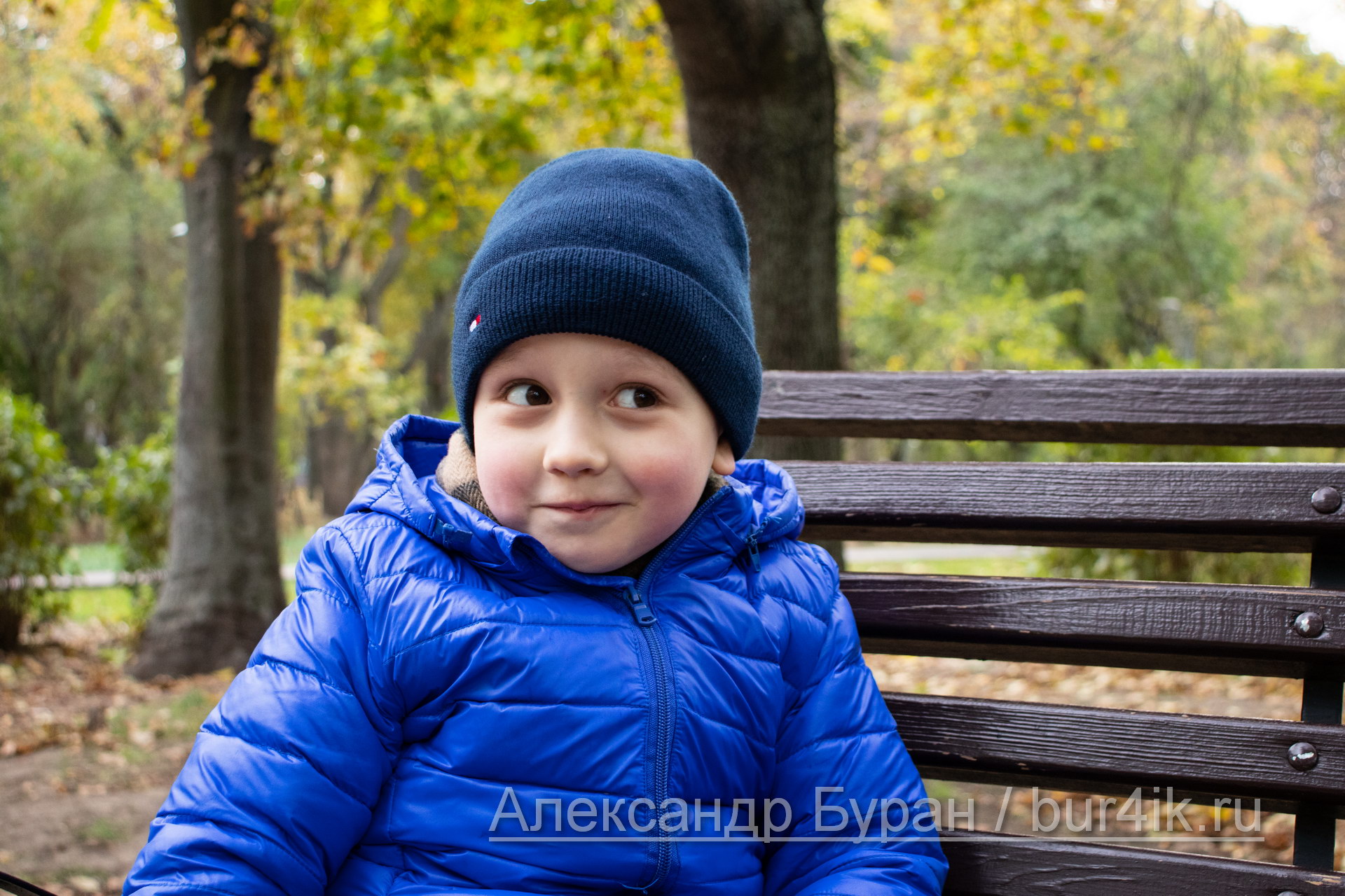 Портрет улыбающегося мальчика, который сидит на скамейке в парке осенью