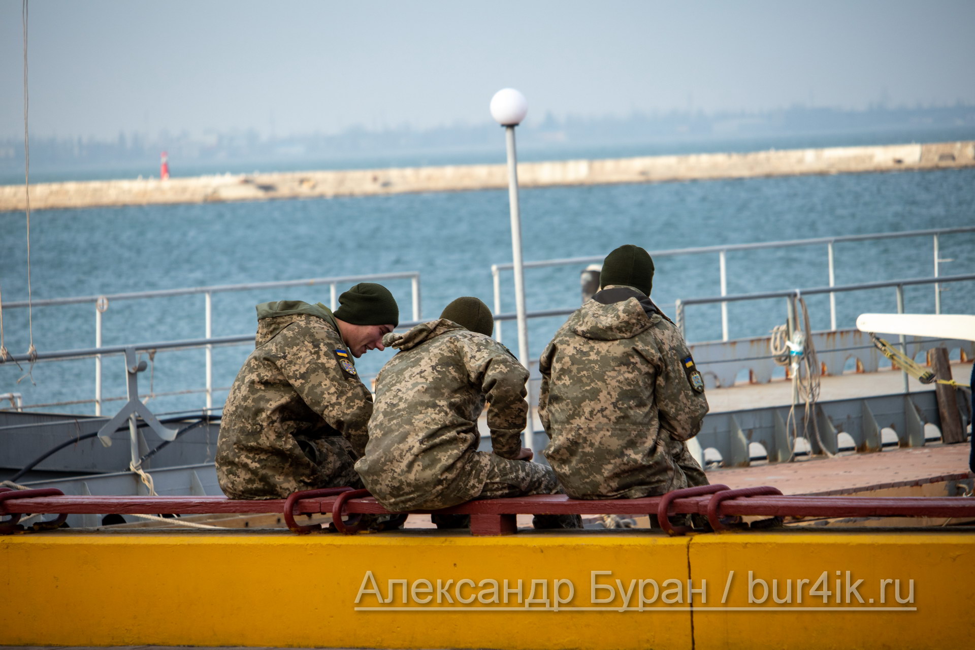 Три солдата в военной форме сидят на скамейке в порту и посмотреть на яхты
