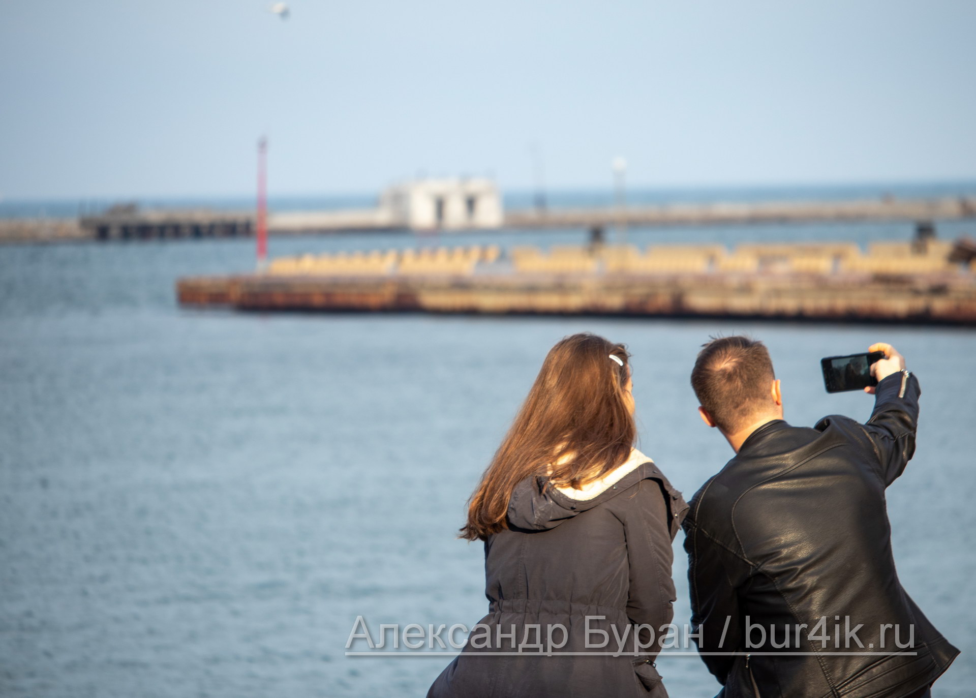 Влюбленная парочка берет селфи, сидя на скамейке в порту, у воды - Украина, Одесса, 09,11,2019