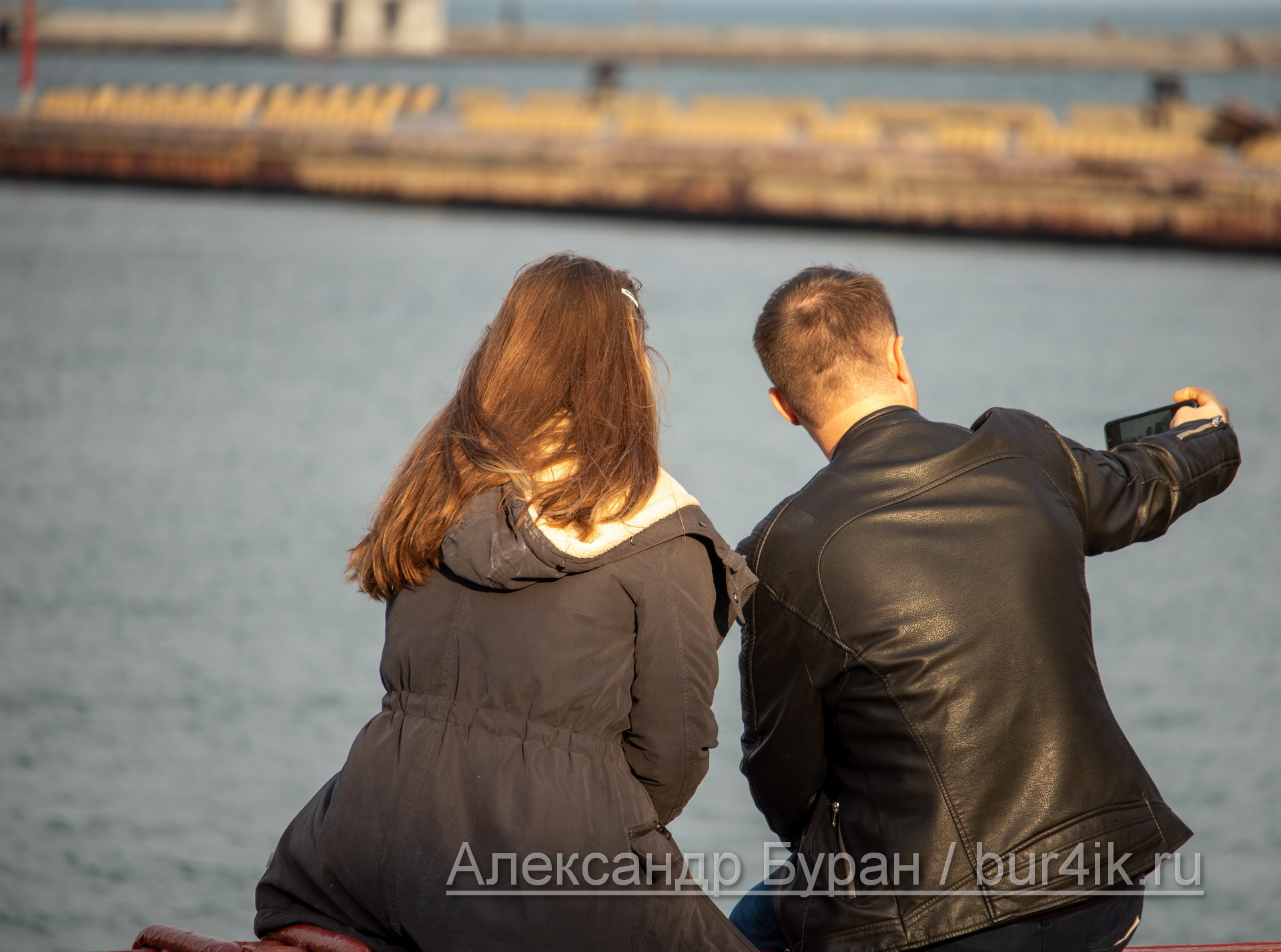 Парень и девушка фотографируют себя на смартфон, сидя у воды в порту