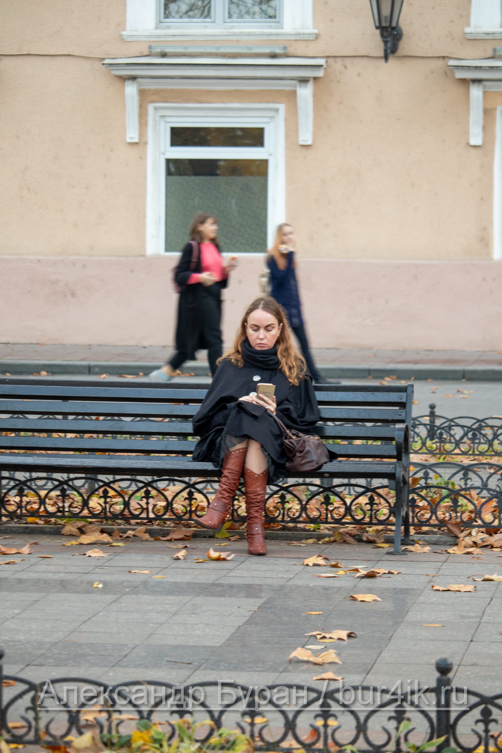 Женщина в черном пальто и коричневые сапоги с телефоном в руках сидит на скамейке в парке - Украина, Одесса, 09,11,2019